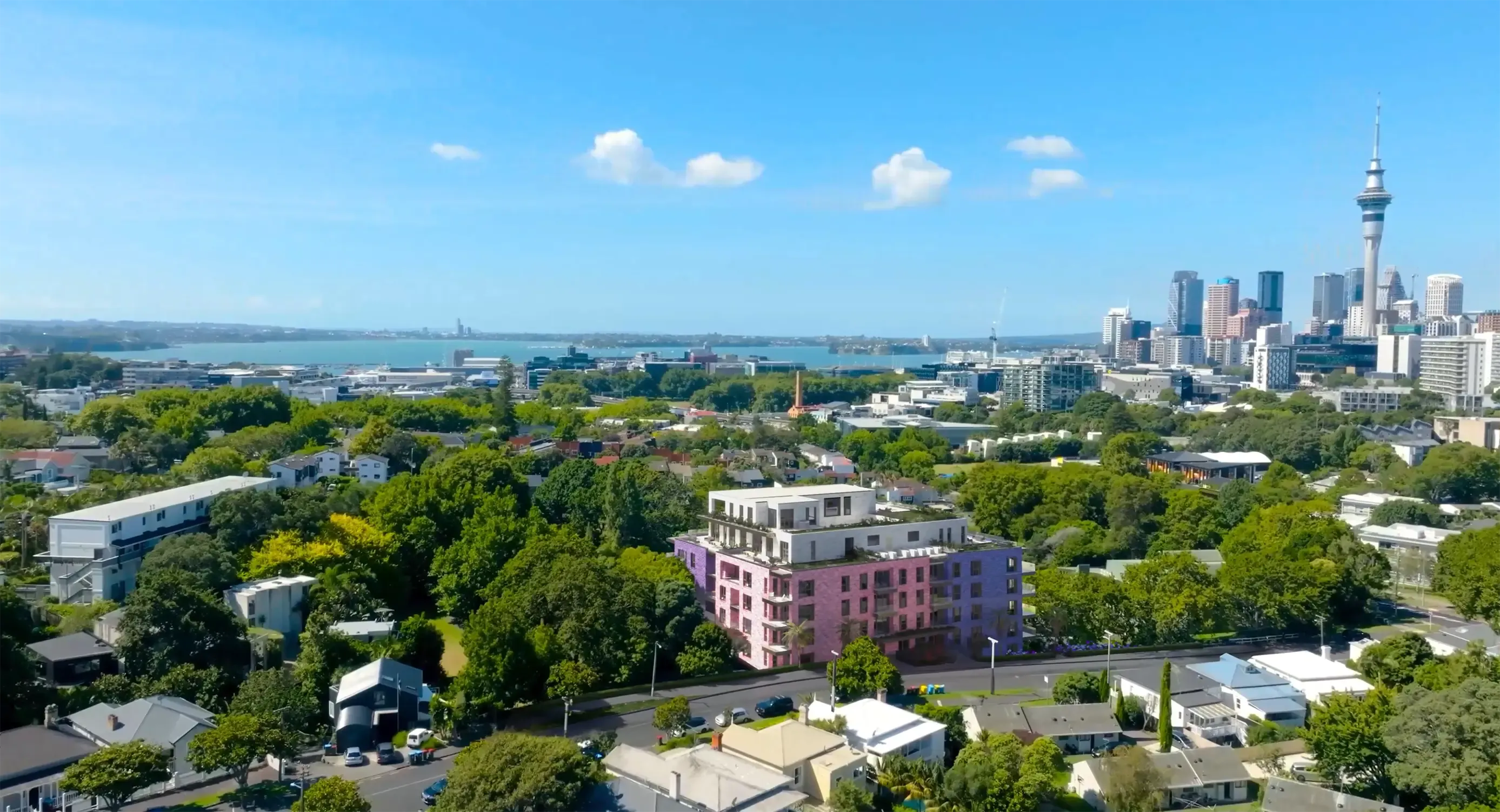 Aerial architectural render of The Kubric, a mid-rise apartment building in Ponsonby, Auckland, featuring pink and purple brick facades with a white rooftop penthouse level and planted terraces. The Auckland CBD skyline and Sky Tower are visible to the right, with the Waitematā Harbour and North Shore stretching beyond.
